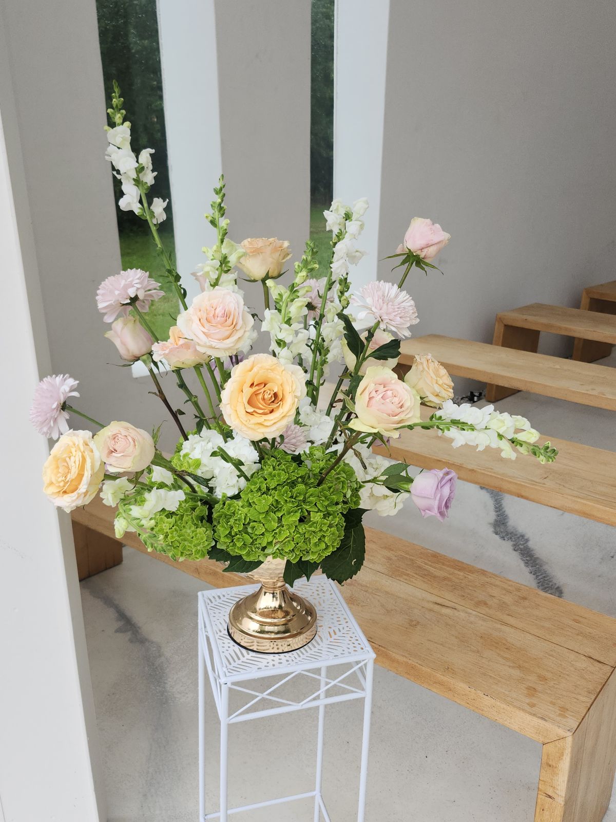 Blush roses, snapdragons, and green hydrangea elevated on a gold stand at the altar — soft ceremony florals that photograph beautifully.