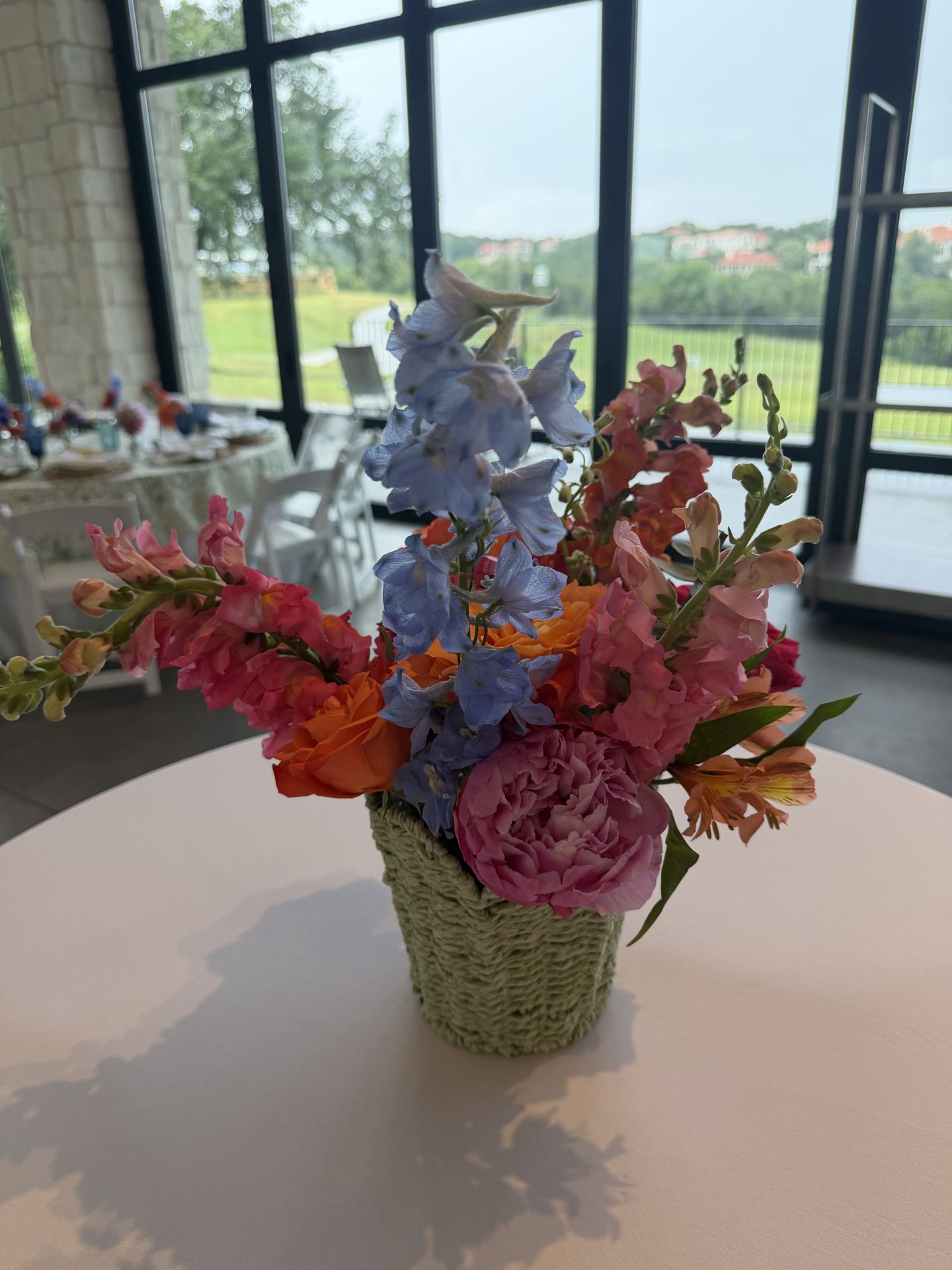 Snapdragons, blue delphinium, peonies, and garden roses gathered in a woven basket — bright, organic reception florals.