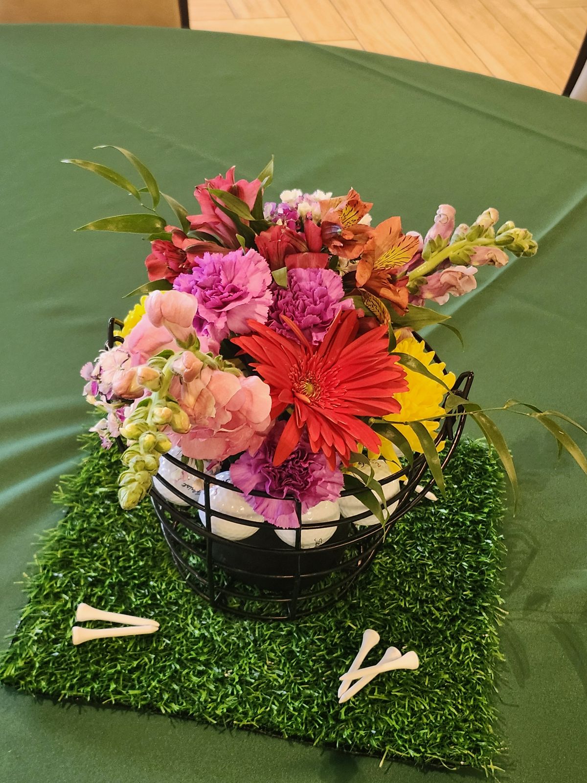 Pink snapdragons and gerberas rising from a basket of golf balls and tees on turf — a centerpiece built for tournament day.