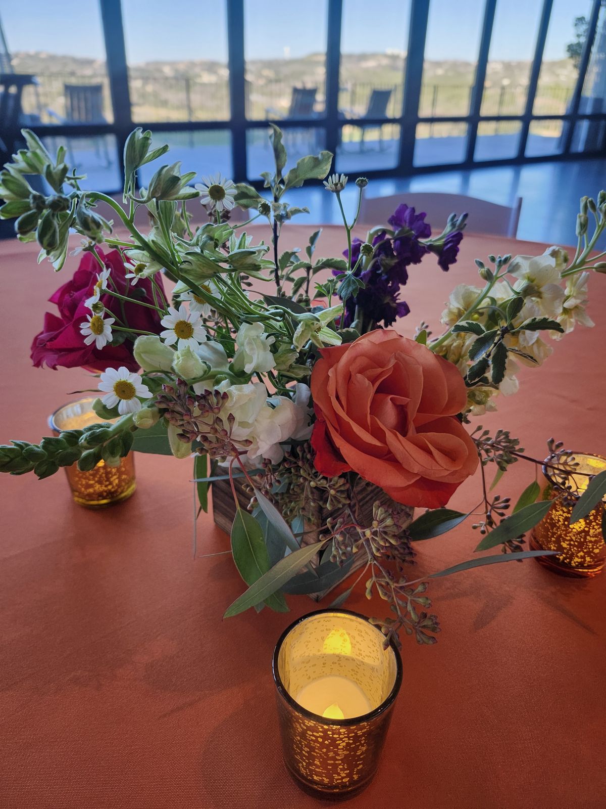 A playful mix of coral roses, daisies, and stock across bud vases and gold votives — set against Hill Country views.