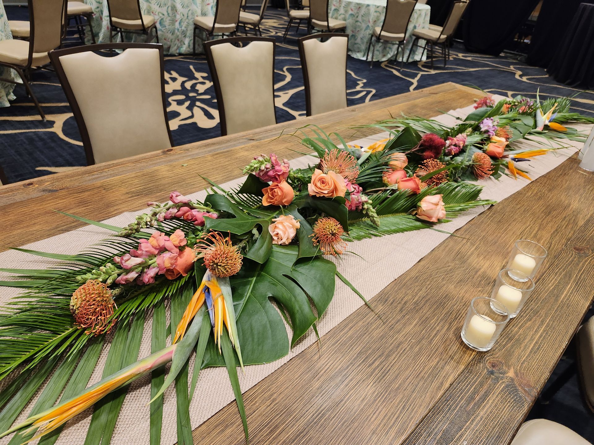 Monstera, palms, birds of paradise, and pincushion protea flowing down a farm table — a lush tropical runner for brand events.